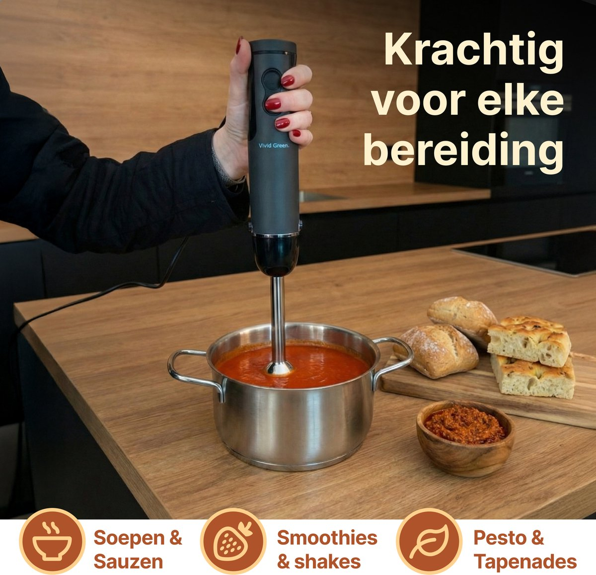 A hand holds an immersion blender in a pot of tomato soup. Bread and pesto are nearby.