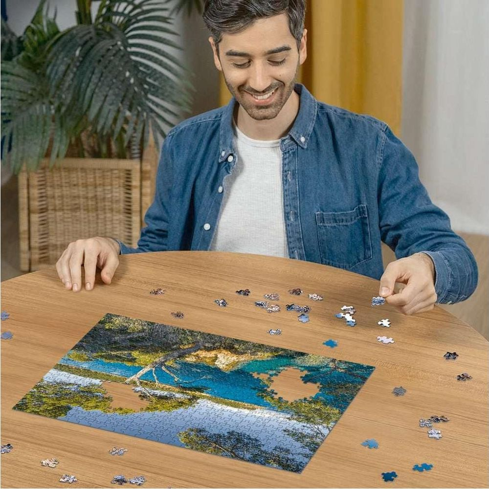Mann baut ein Puzzle eines Sees mit Bäumen und Himmel. Blauer Himmel, Wasser, grüne Bäume.