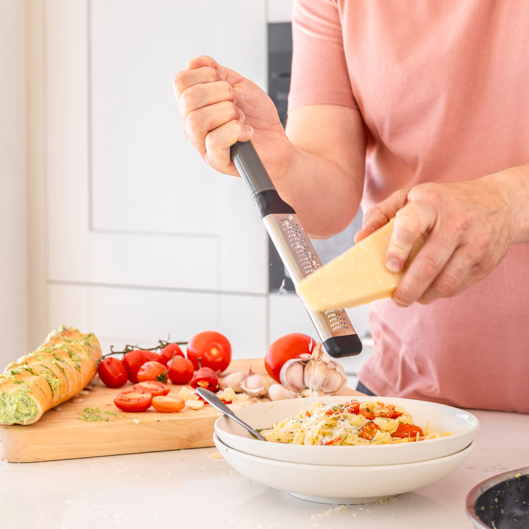 Eine Person reibt Parmesan über ein Nudelgericht, daneben Brot, Tomaten und Knoblauch.