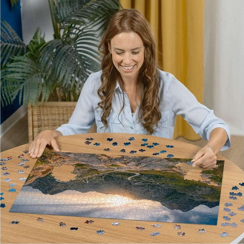 Frau löst ein Puzzle mit Berglandschaft bei Sonnenuntergang auf einem Holztisch.