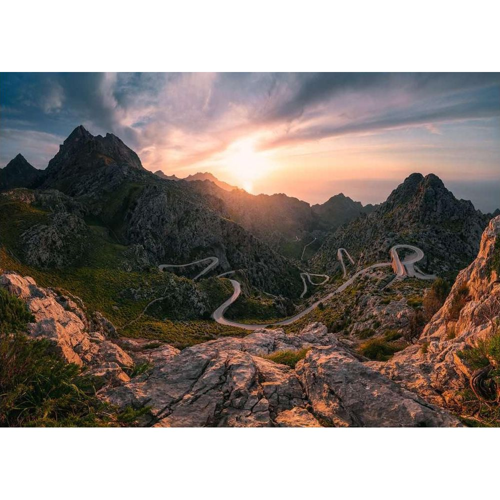 Serpentinenstraße in bergiger Landschaft bei Sonnenuntergang, Felsen im Vordergrund.