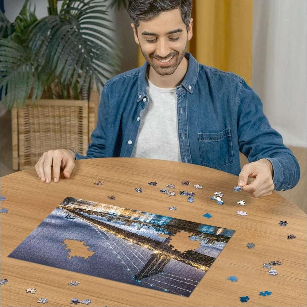 Mann zusammensetzen eines Puzzles einer Brücke bei Nacht, mit Lichtern, die sich im Wasser spiegeln.