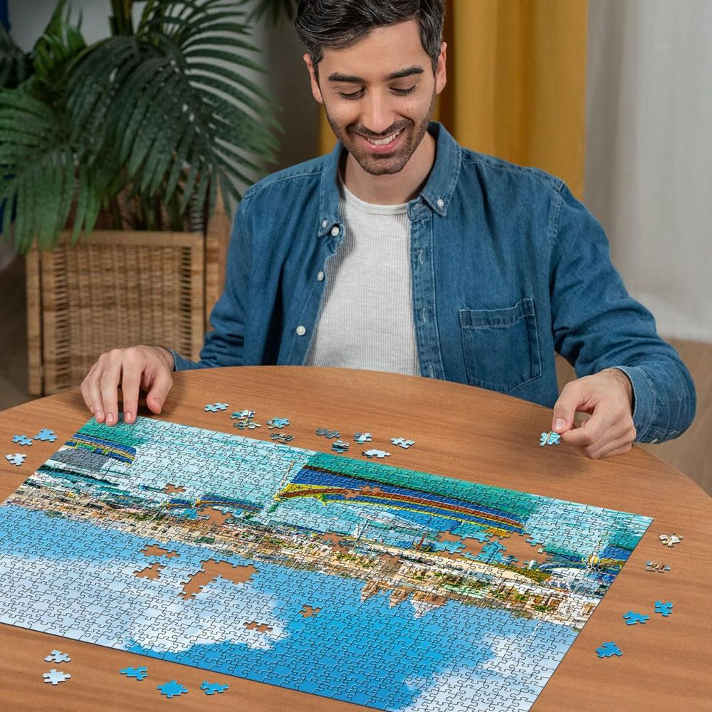 Ein Mann sitzt an einem Holztisch und setzt ein Puzzle zusammen, das eine Stadt am Wasser zeigt.