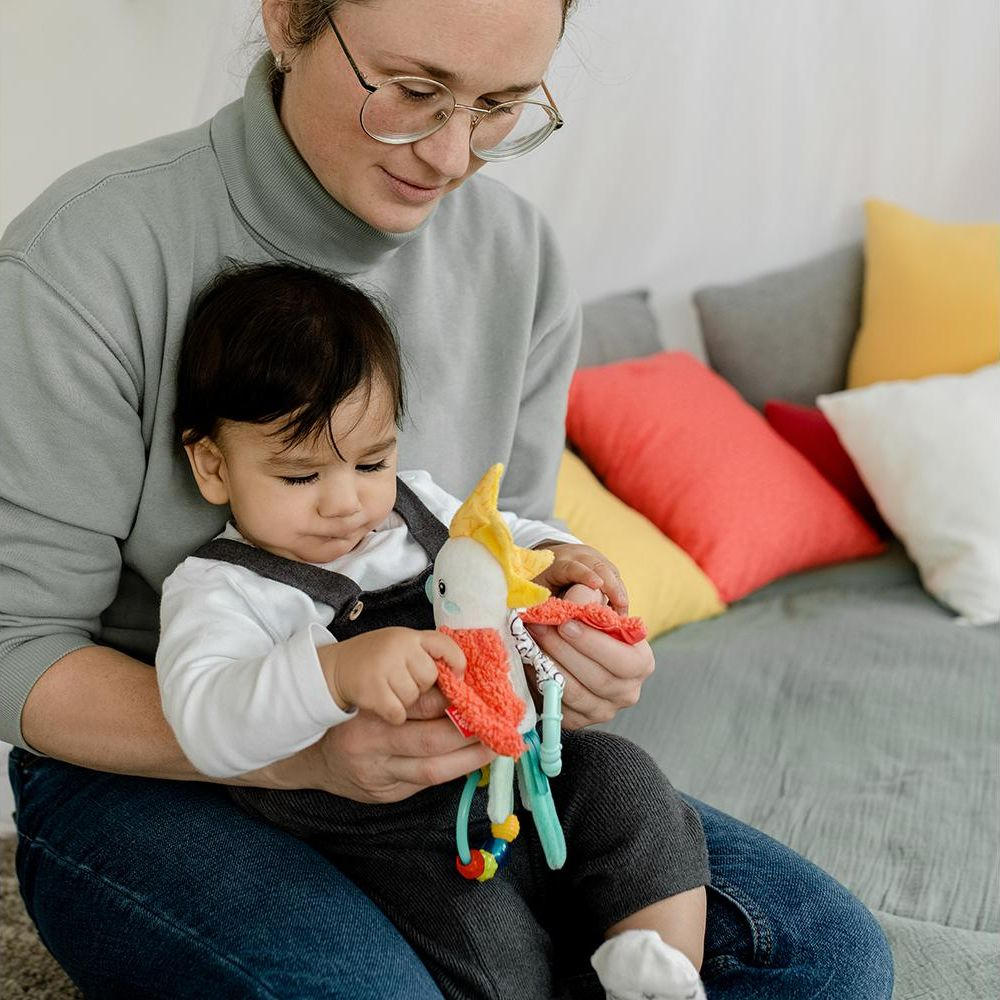 Une femme avec des lunettes tient un bébé. Le bébé joue avec un jouet en peluche coloré.