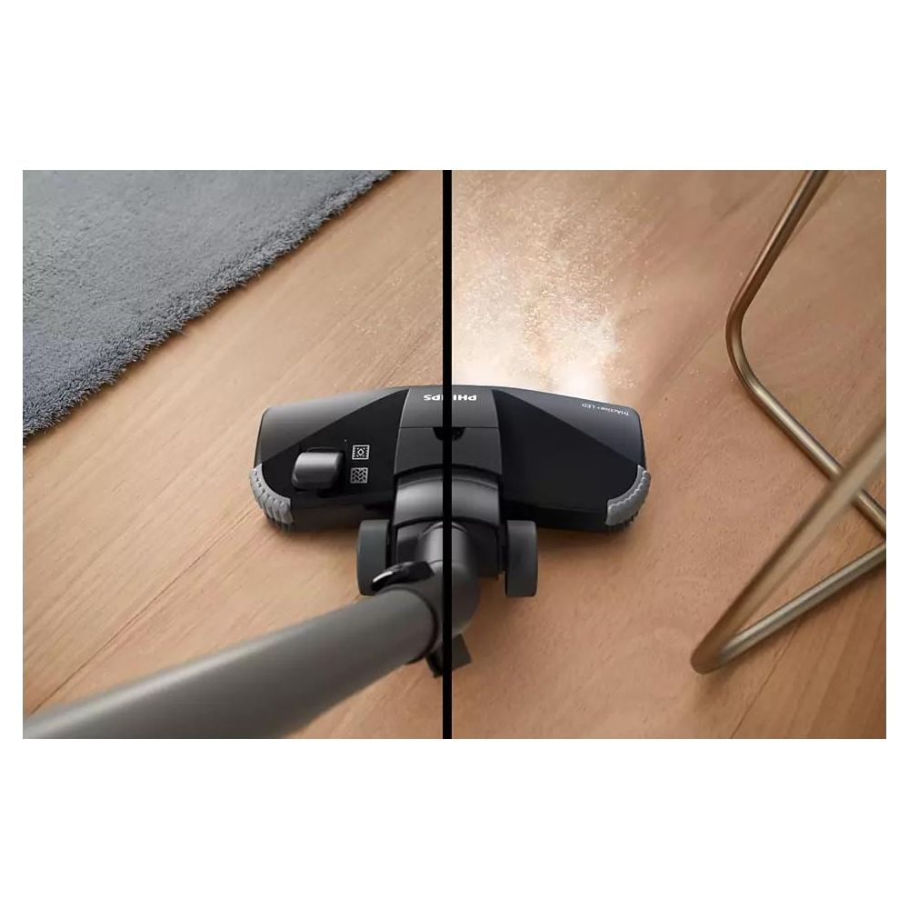Tête d'aspirateur noire nettoyant un parquet. Tapis à gauche, poussière soulevée à droite.