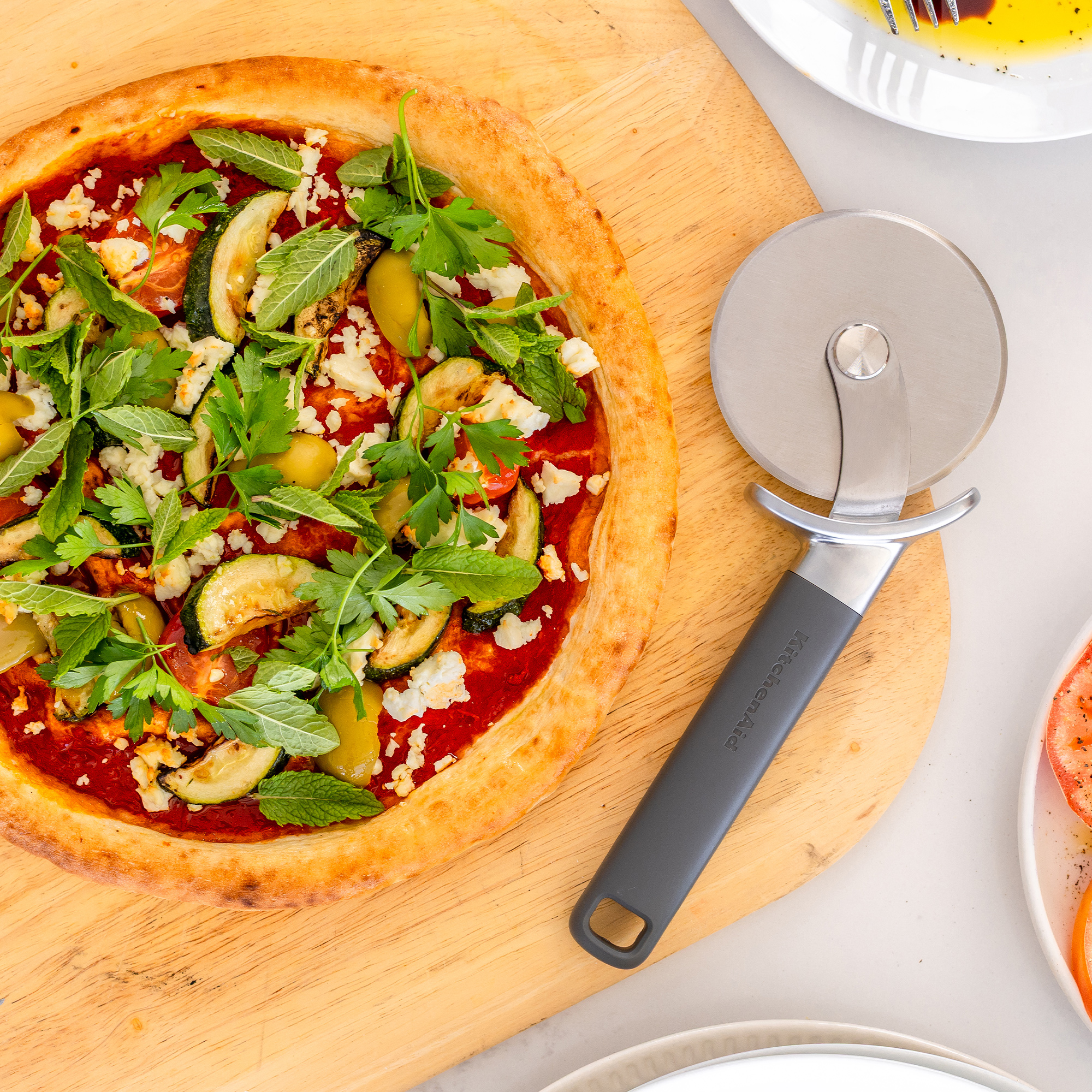 Eine Pizza mit Zucchini, Oliven, Käse und Kräutern auf einem Holzbrett mit Pizzaschneider.