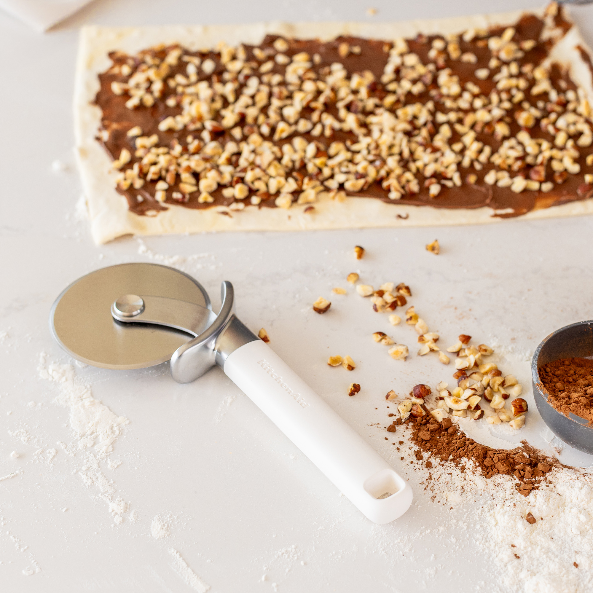 Ein Teig mit Schokoladencreme und Nüssen, daneben ein Pizzaschneider und Kakaopulver.