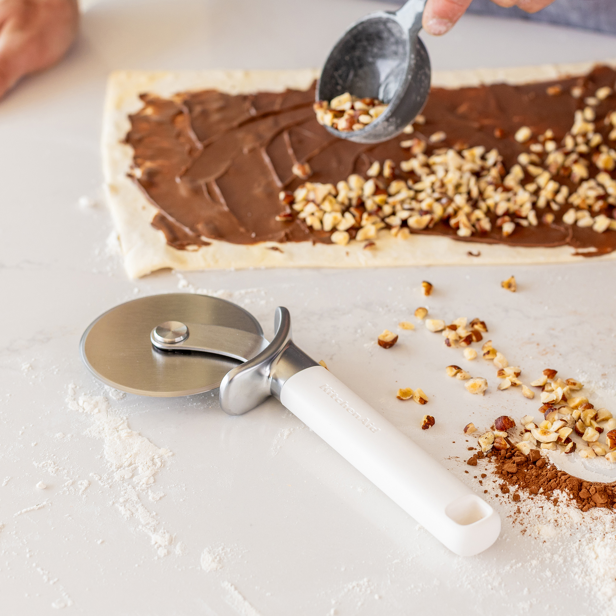 Nüsse werden auf Schokoladencreme auf Teig gestreut. Ein Pizzaschneider liegt daneben.