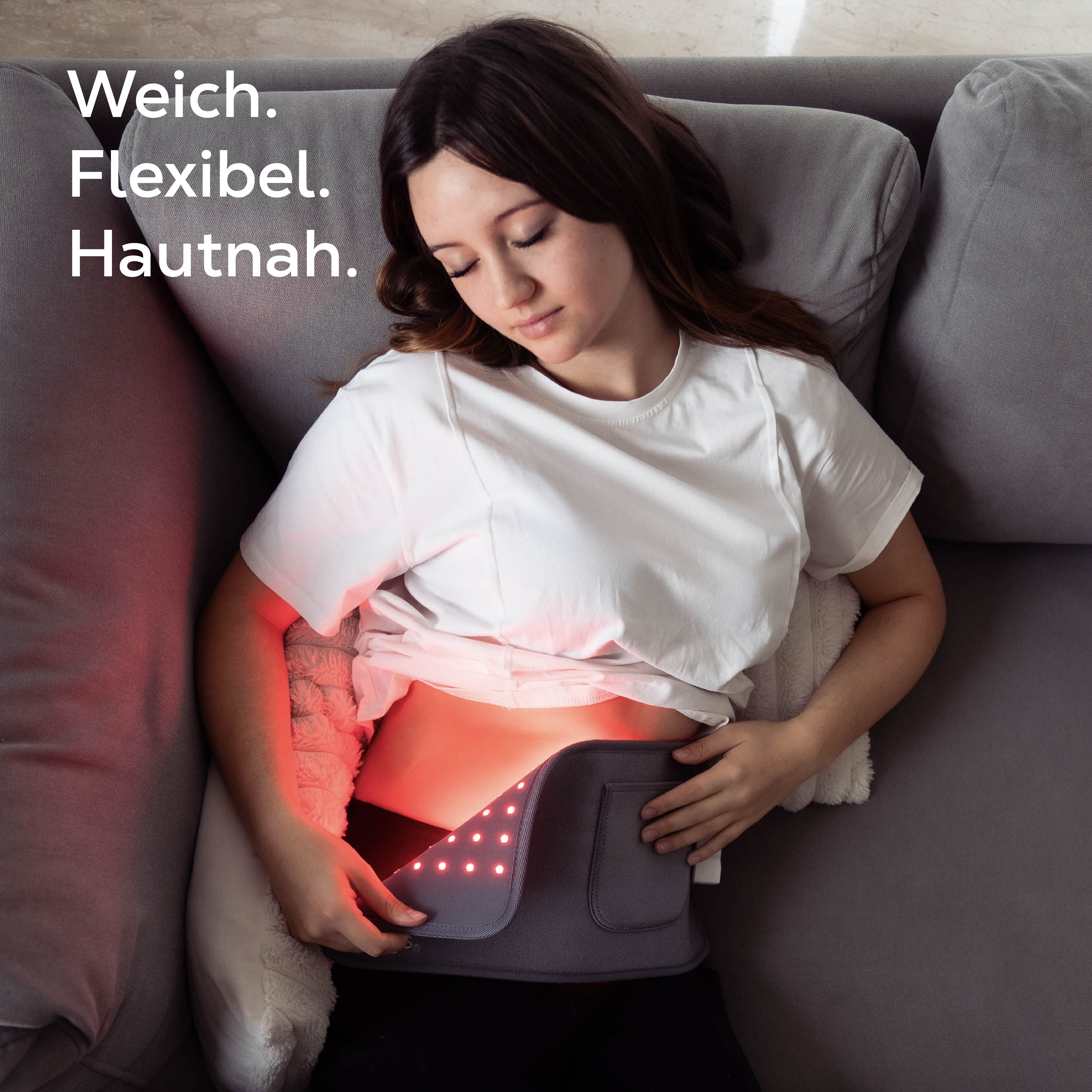 Junge Frau liegt auf Sofa mit roter Lichttherapie-Gürtel am Bauch.
