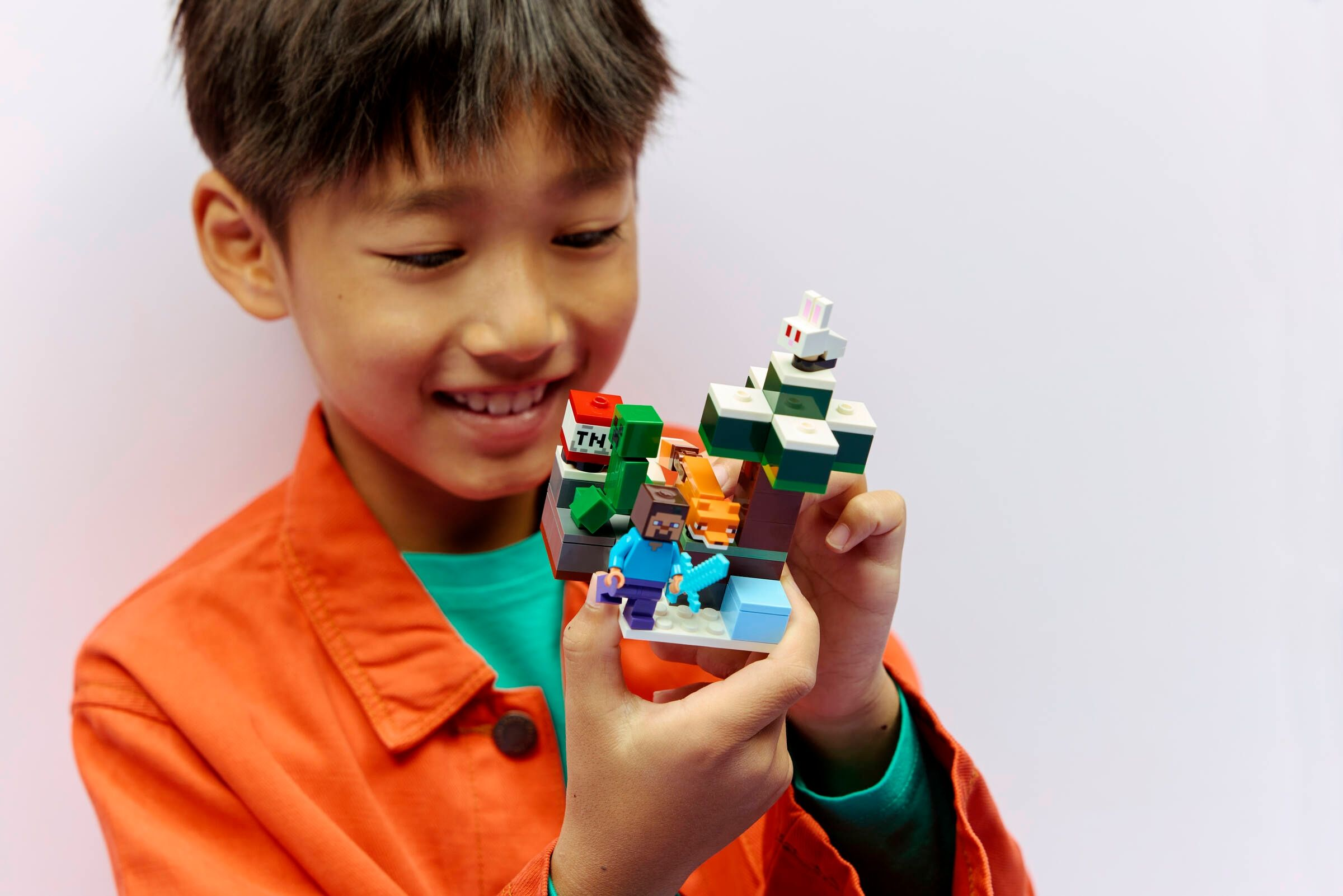 Ein Junge in einem orangen Hemd hält ein Minecraft-LEGO-Set mit Figuren und einem Fuchs.