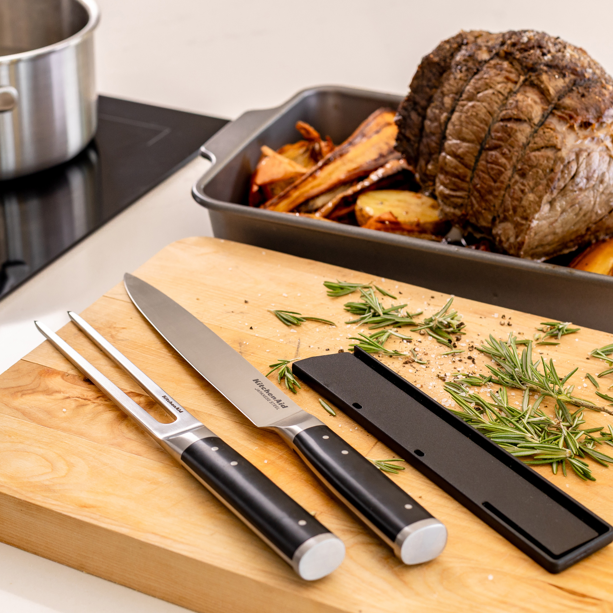 Ein Braten mit Kartoffeln und Karotten im Backblech, daneben ein Fleischgabel- und Messerset.