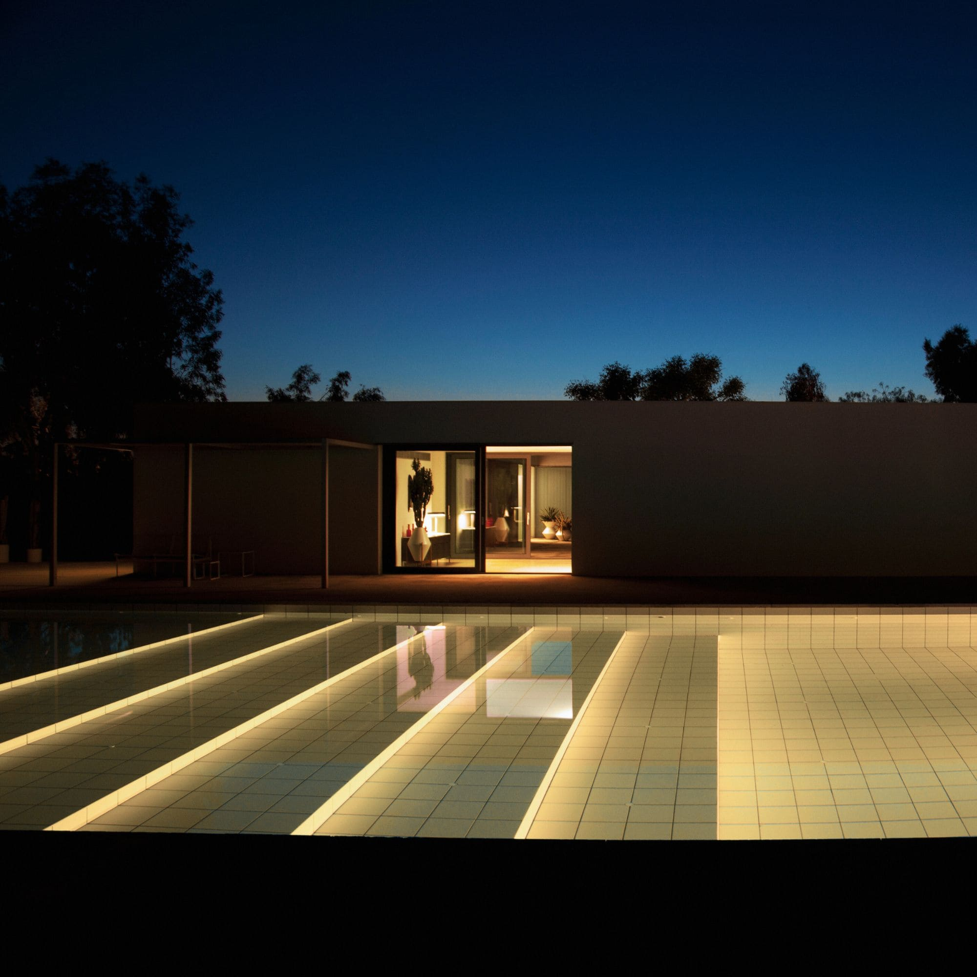 Casa moderna de noche con piscina iluminada y luces interiores visibles.