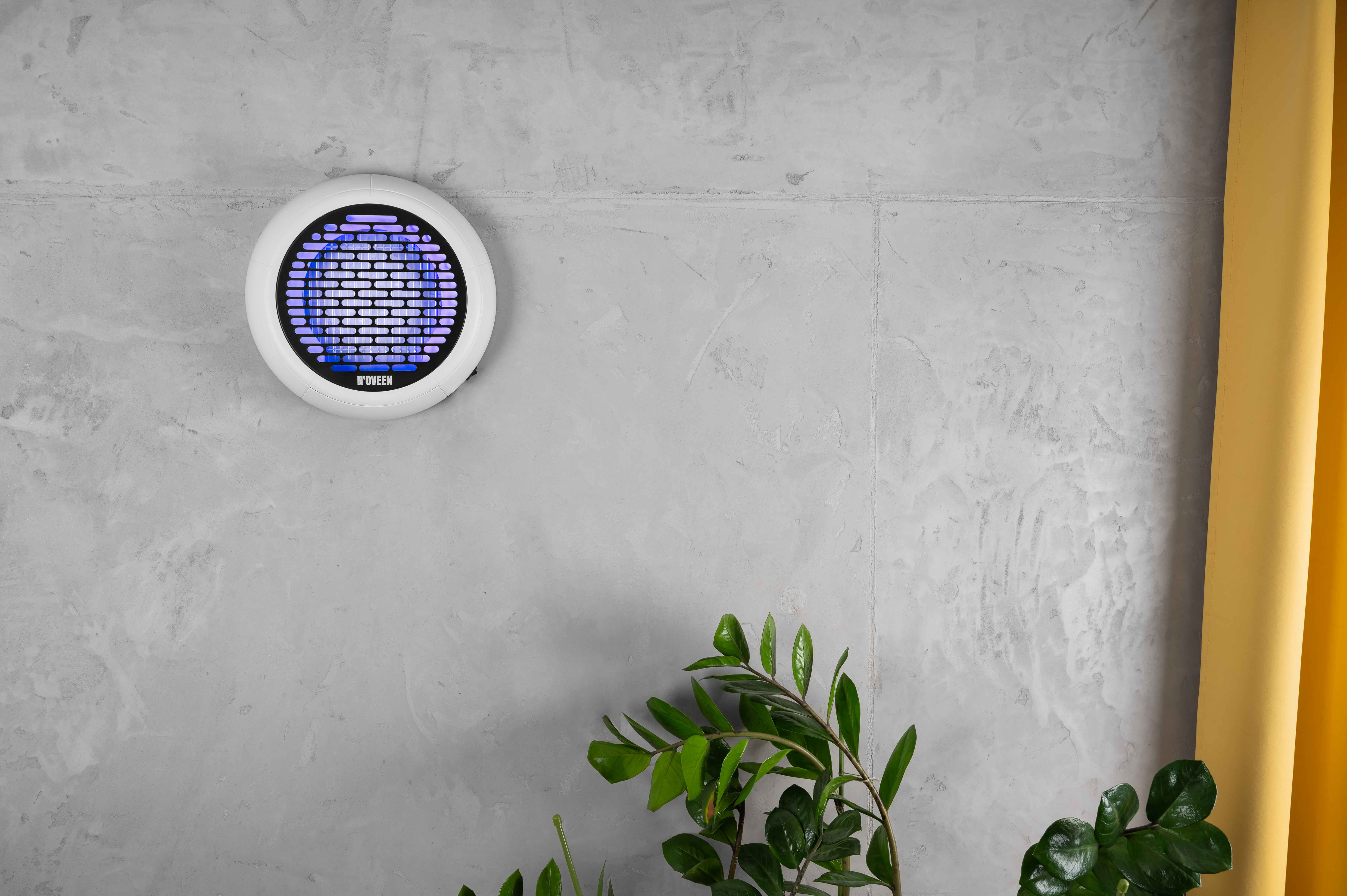 Biała lampa owadobójcza z niebieskim światłem UV wisi na betonowej ścianie obok zielonej rośliny.