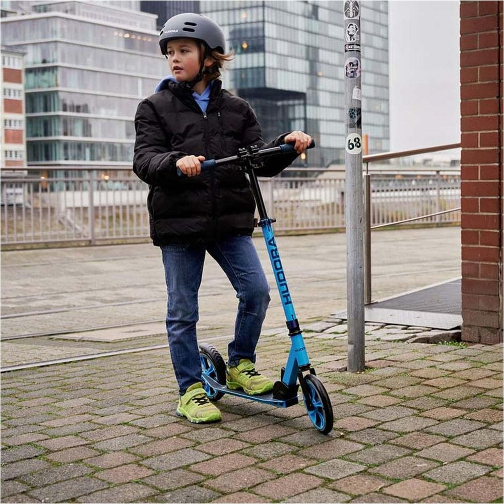 Ein Junge mit Helm steht auf einem blauen Roller auf einem gepflasterten Weg.