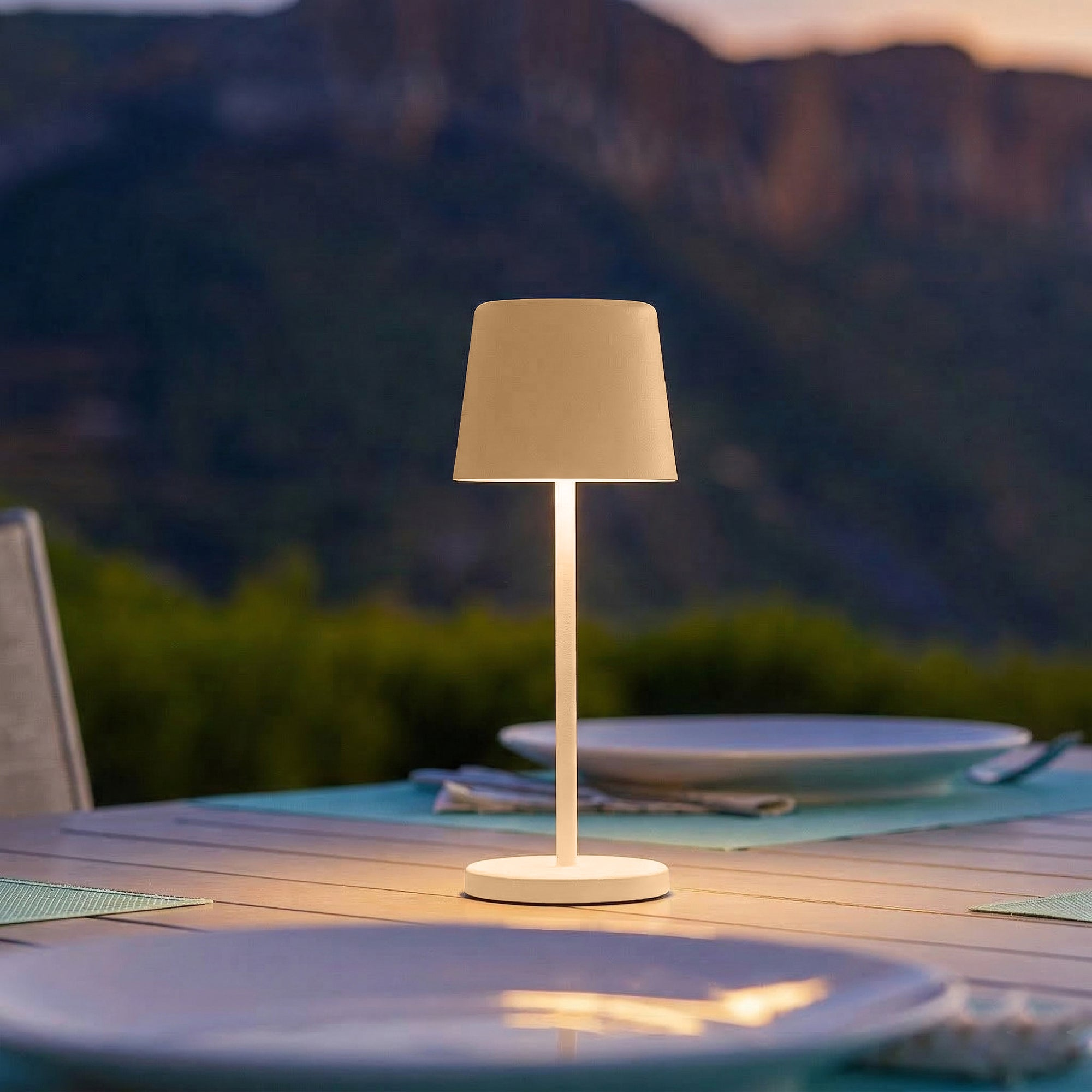 Eine Tischlampe leuchtet auf einem Holztisch im Freien mit Bergkulisse.