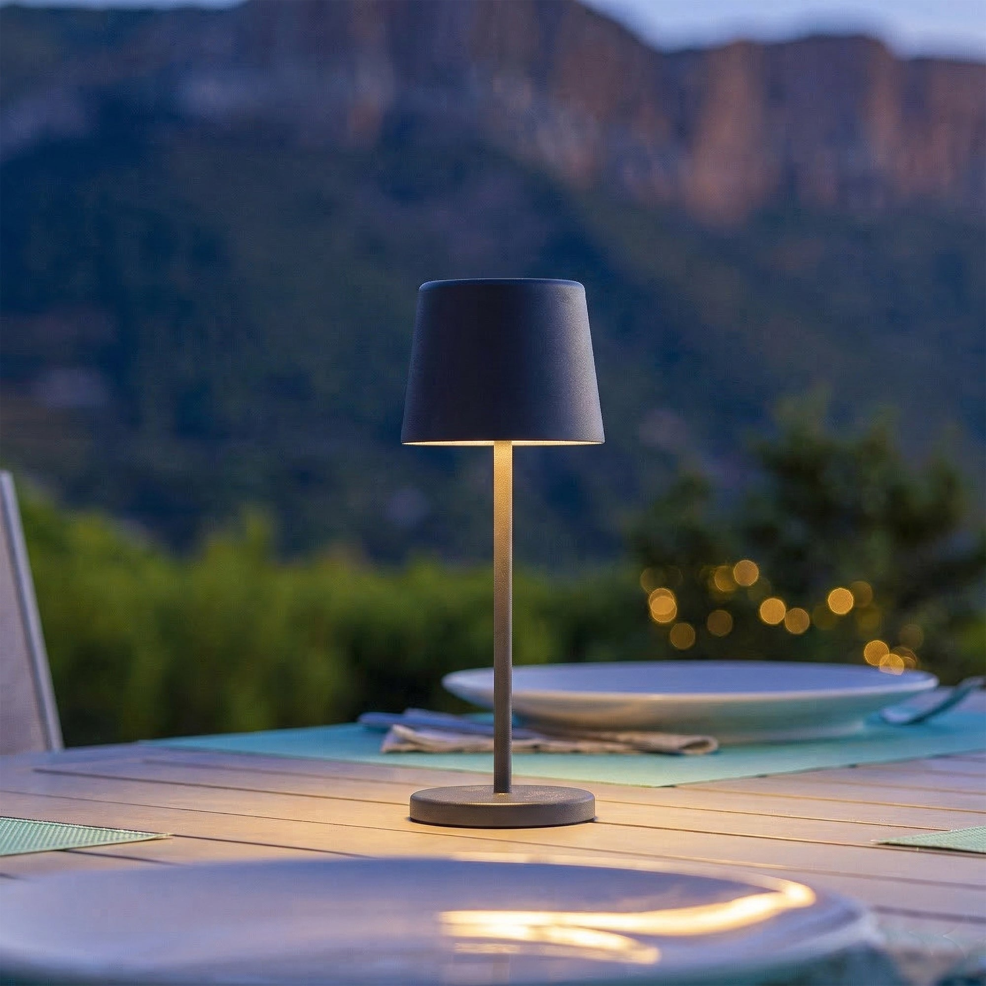 Eine dunkle Tischlampe steht auf einem Holztisch im Freien, mit Bergen im Hintergrund.
