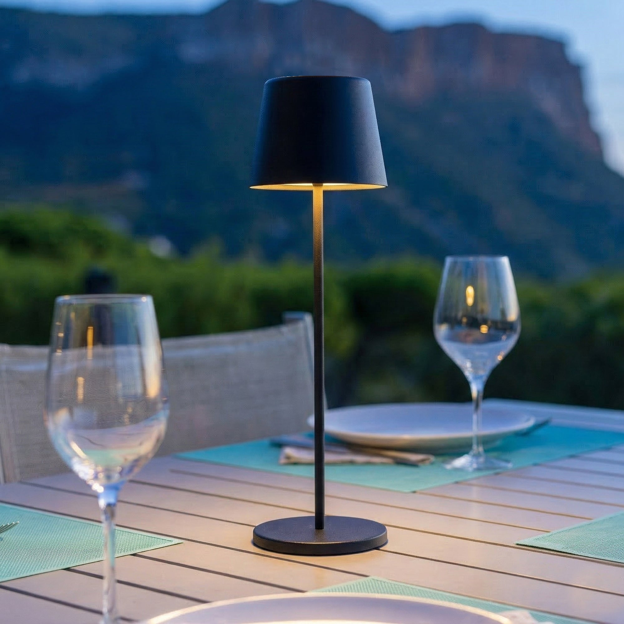 Eine schwarze Tischlampe leuchtet auf einem Gartentisch mit Gläsern und Bergen im Hintergrund.