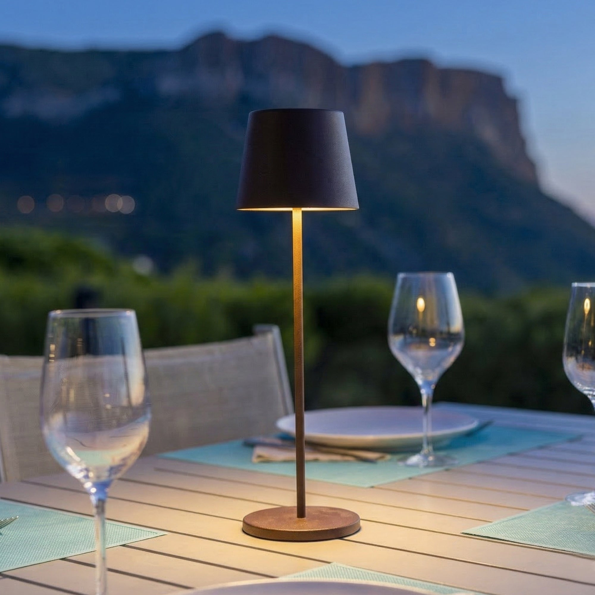 Ein Tisch mit Gläsern, Tellern und Lampe im Freien bei Dämmerung mit Bergen im Hintergrund.