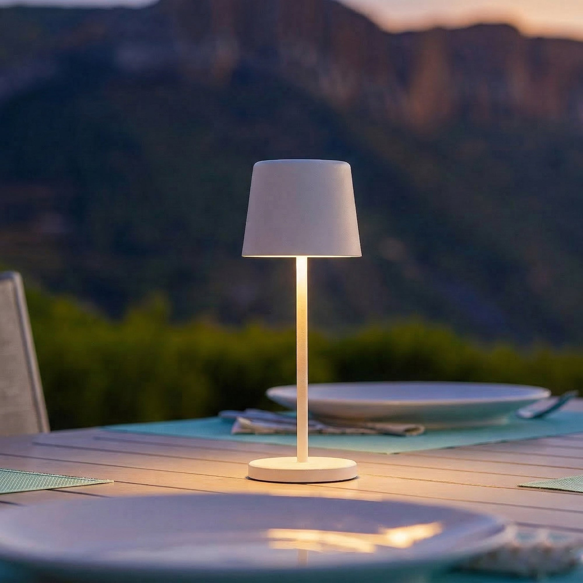 Eine weiße Lampe steht auf einem Tisch im Freien vor einem Berg und Bäumen.