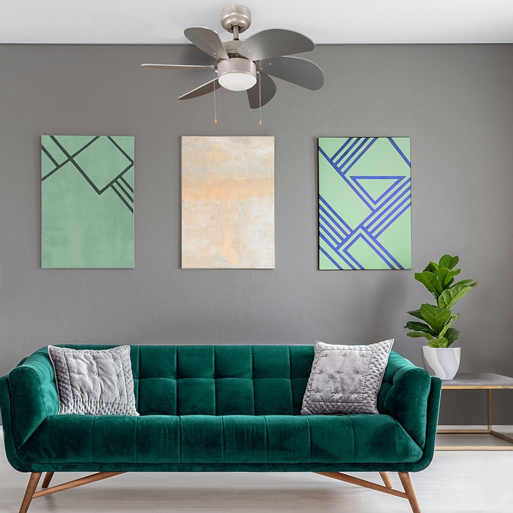 Un sofá de terciopelo verde frente a una pared gris con tres cuadros y un ventilador.