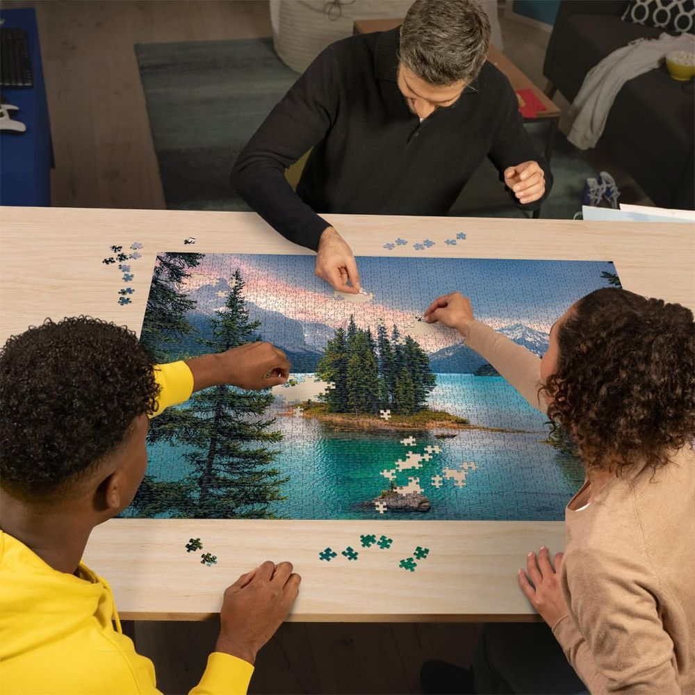 Tre persone assemblano un puzzle di un paesaggio montano e lacustre su un tavolo.