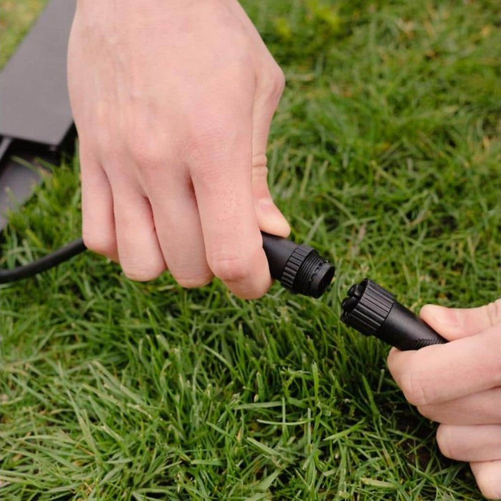 Zwei Hände verbinden schwarze Kabelstecker über grünem Gras.