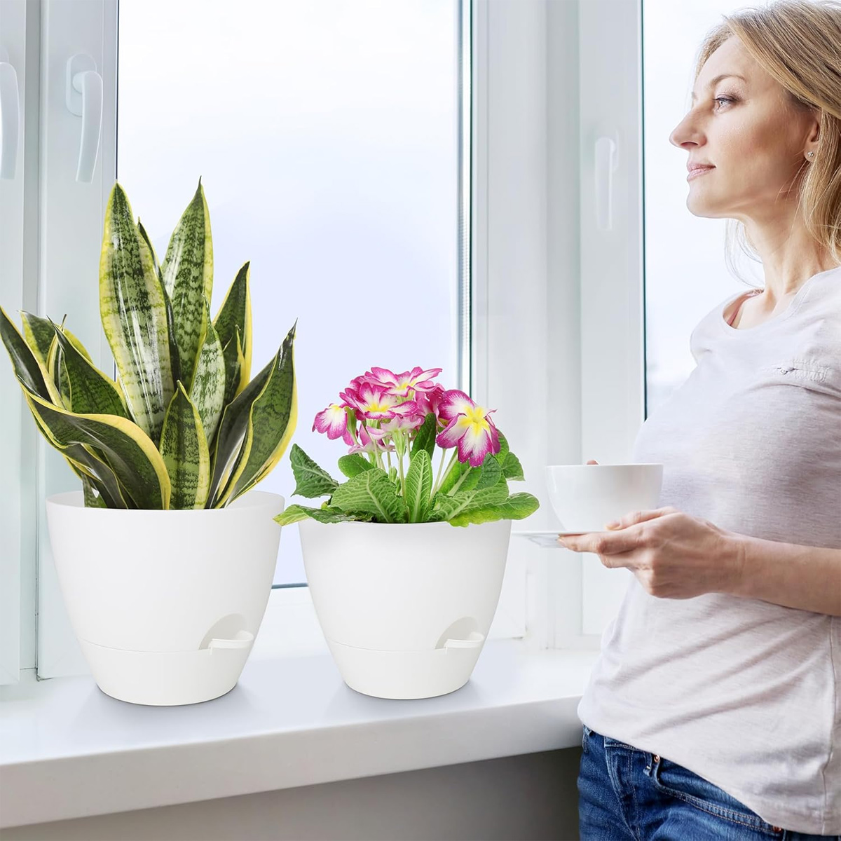 Frau mit Tasse blickt aus dem Fenster auf Bogenhanf und pinke Blumen auf der Fensterbank.