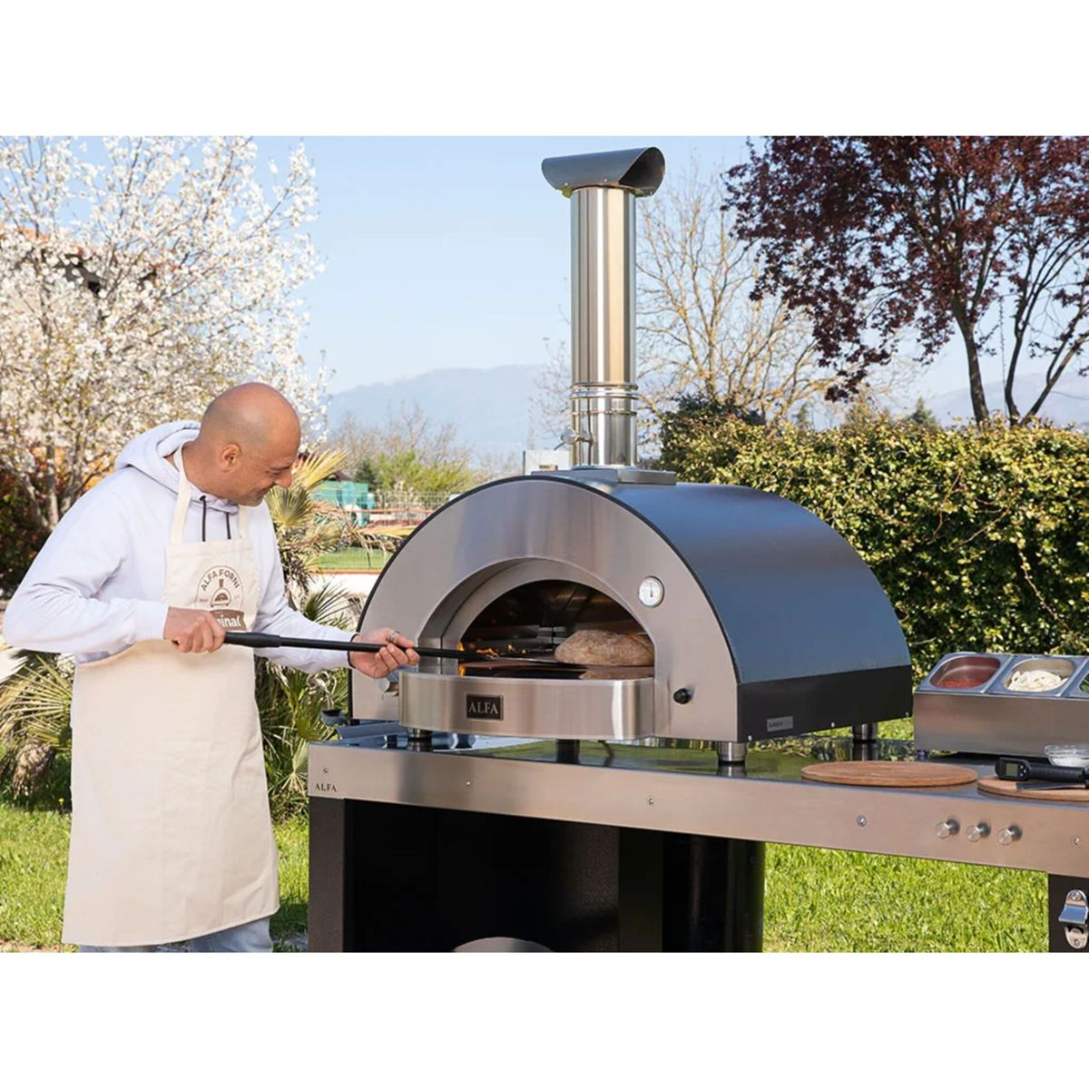 Ein Mann mit Schürze benutzt einen Pizzaschaufel in einem Ofen im Freien.