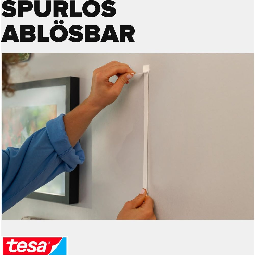 Una mano stacca una striscia bianca da un muro, con un quadro incorniciato a sinistra.