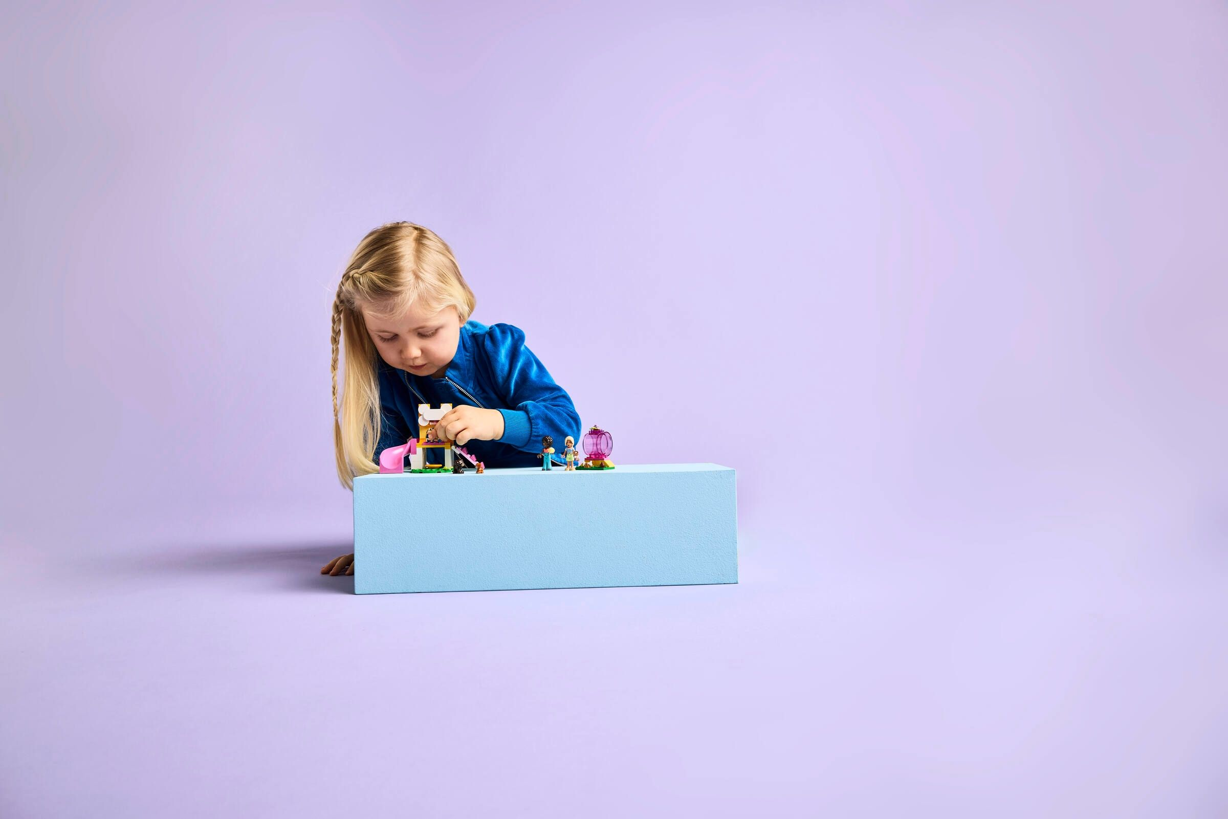 Ein kleines Mädchen mit blonden Zöpfen spielt mit Lego auf einem blauen Podest.