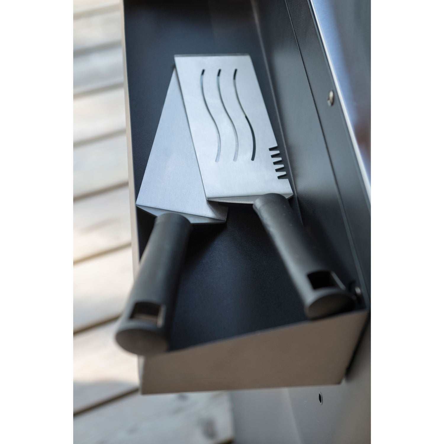 Two metal grill spatulas with black handles rest on a black shelf.