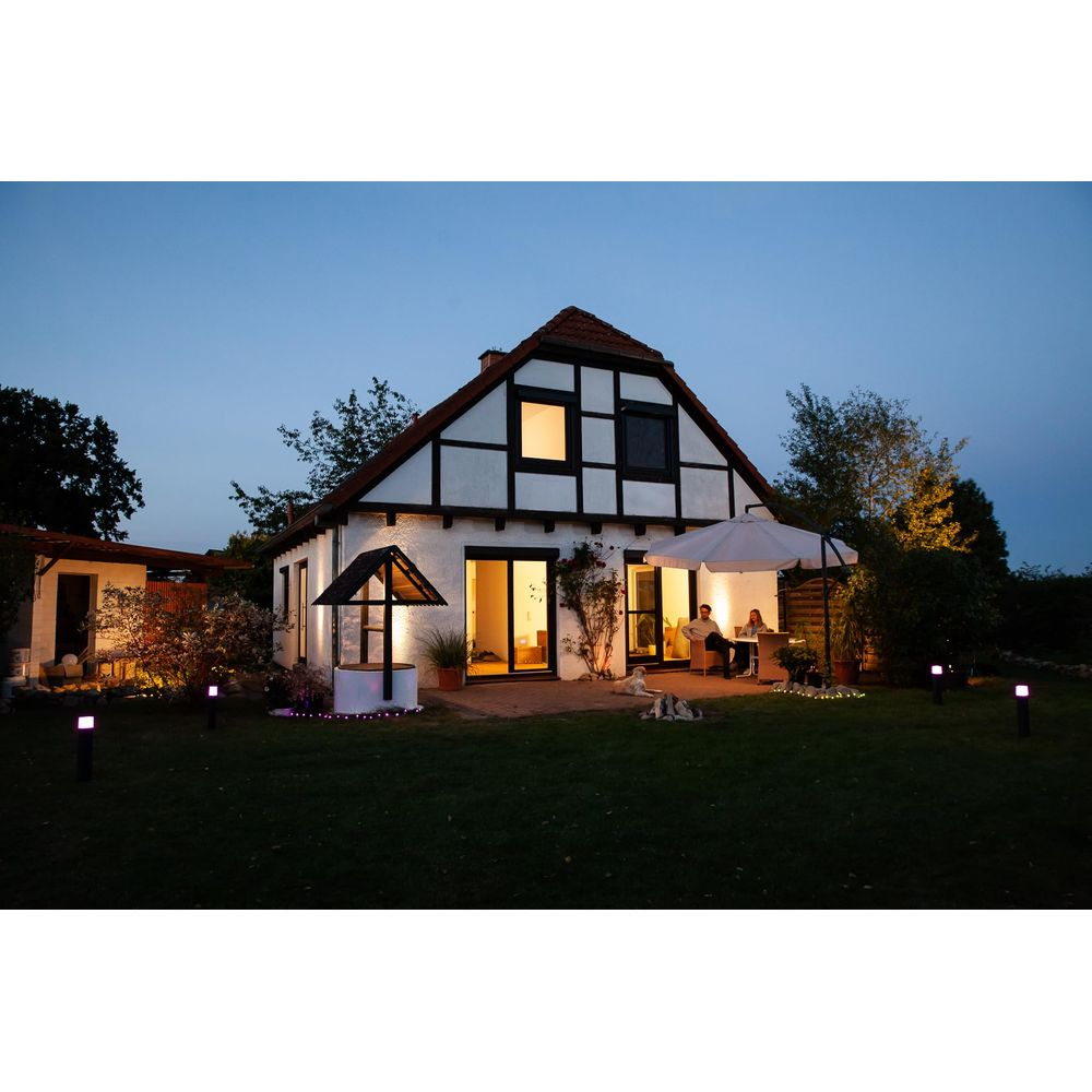 Une maison à colombages la nuit, avec un jardin éclairé et des personnes sur la terrasse.