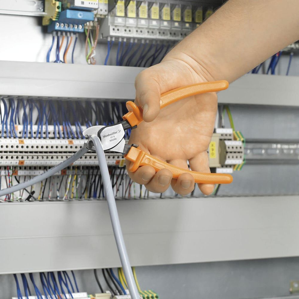 Una mano tiene una pinza arancione che taglia un cavo grigio vicino a un pannello di controllo elettrico.