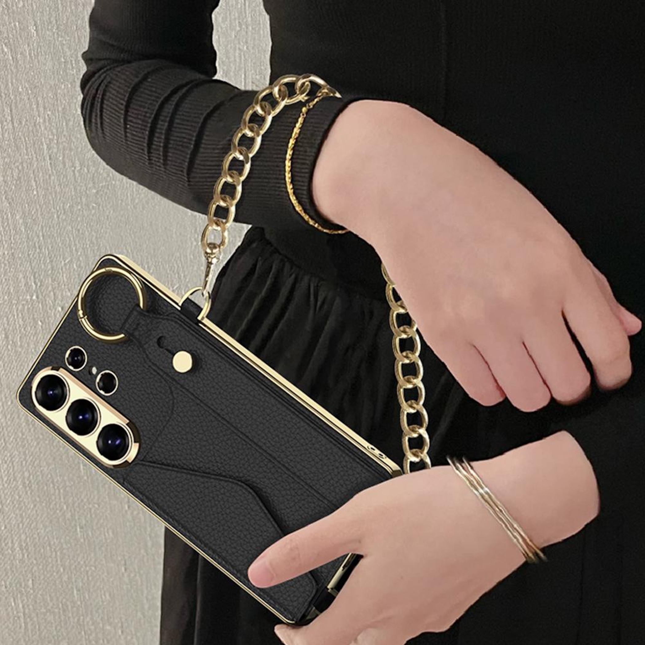 Schwarzes Handy-Etui mit goldener Kette und Ringen, von einer Person in Schwarz gehalten.