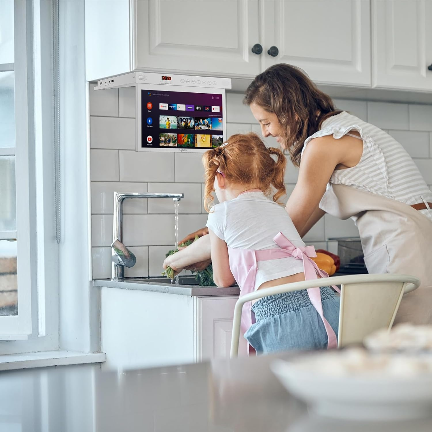 Madre e hija lavan verduras en una cocina, una pantalla cuelga sobre el fregadero.