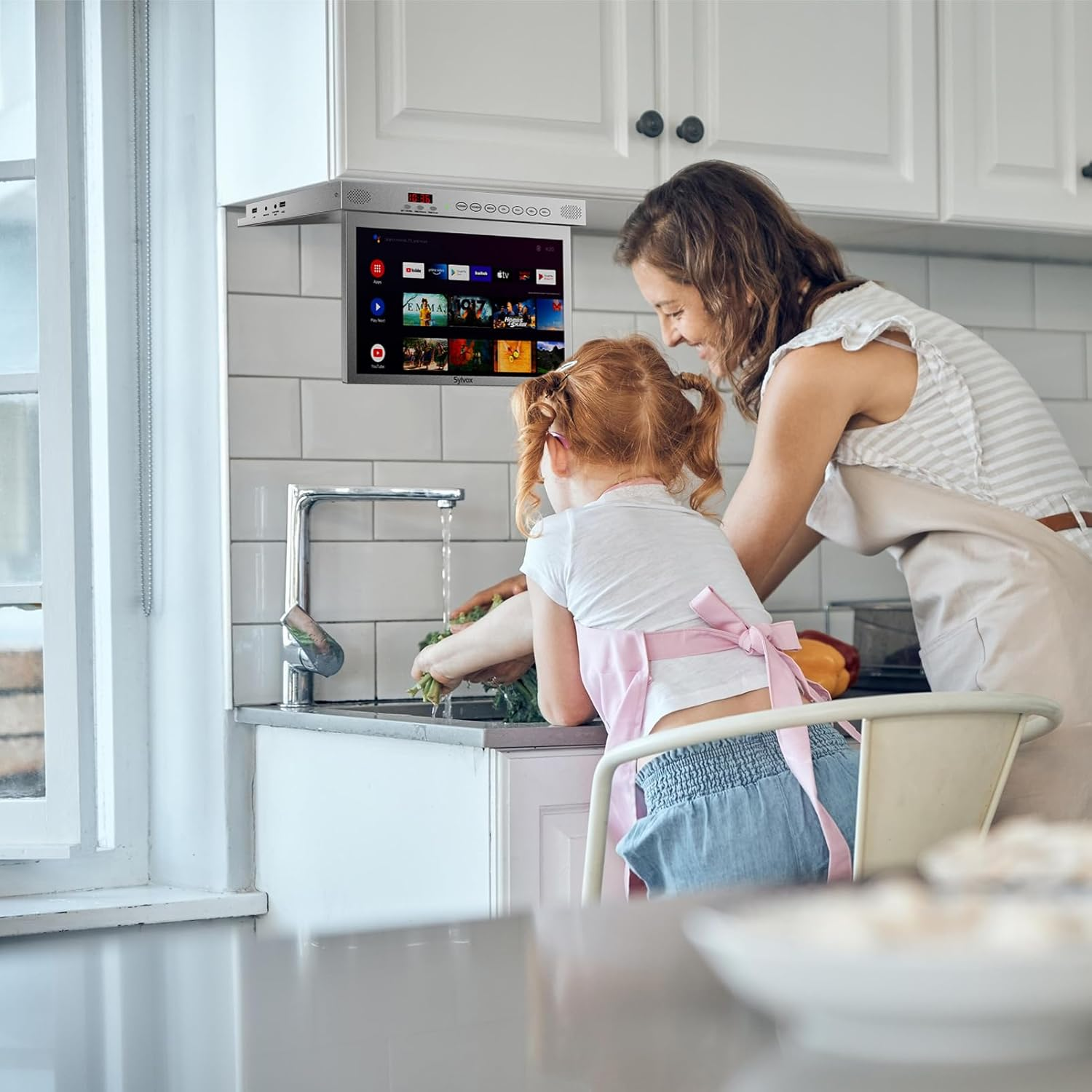 Madre e hija lavando verduras en una cocina bajo una pantalla.