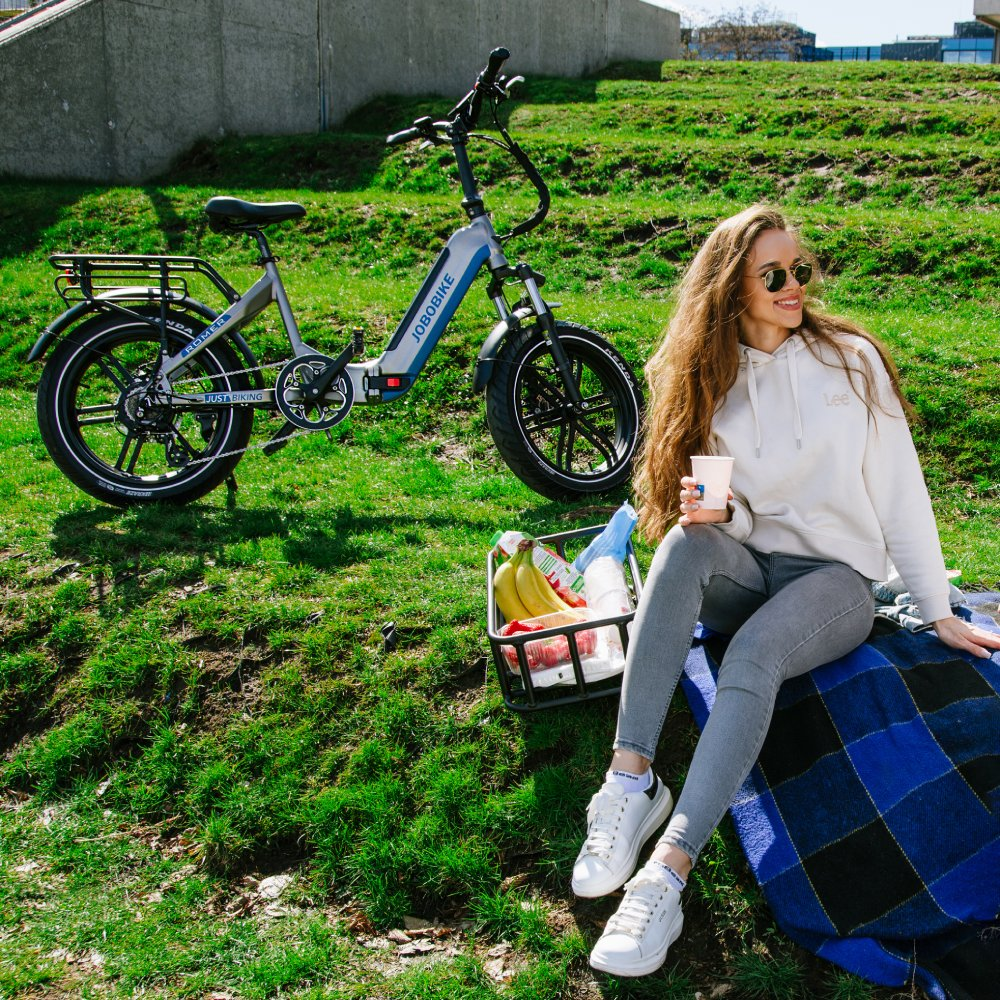 Frau sitzt auf Decke neben Fahrrad mit Picknickkorb auf grünem Hügel.