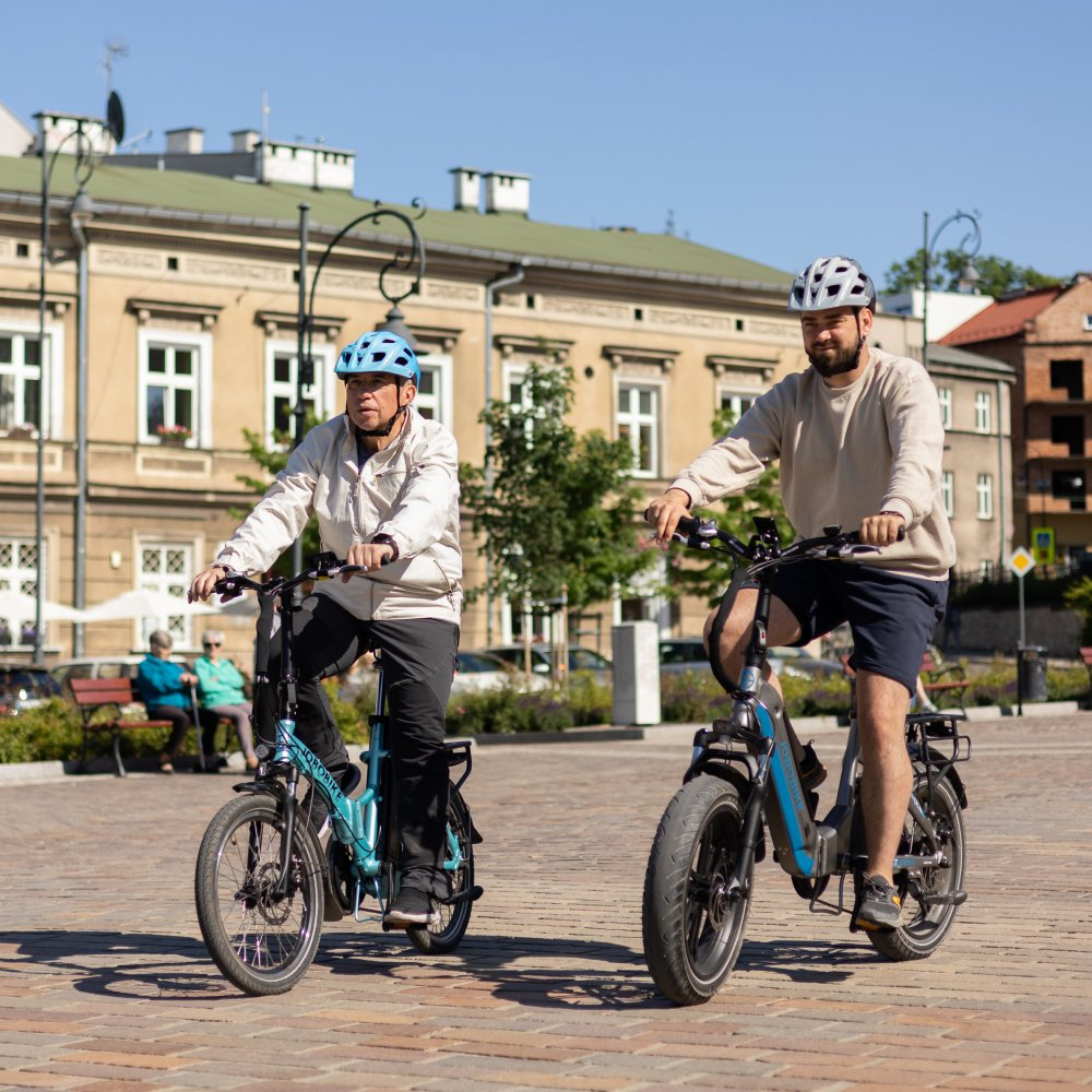 Zwei Männer fahren E-Bikes in einer städtischen Umgebung mit Gebäuden im Hintergrund.