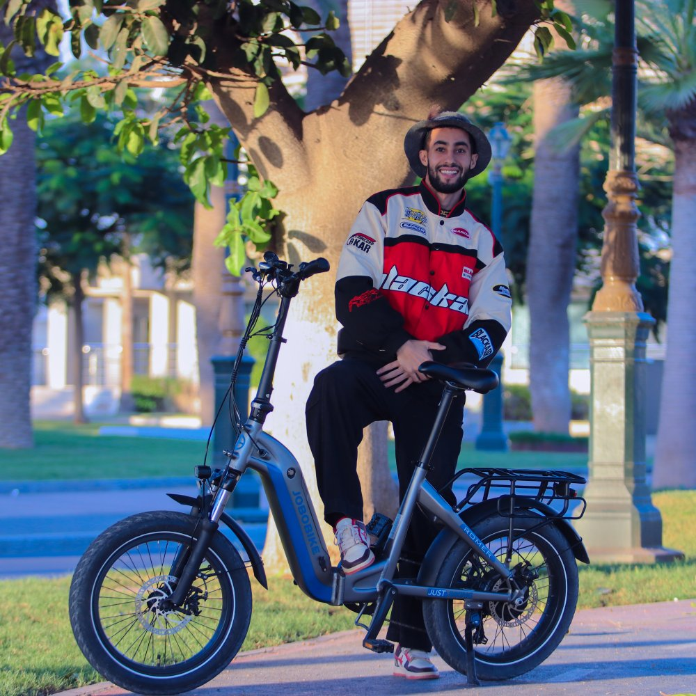 Ein Mann in einem bunten Jackett und Hut sitzt auf einem E-Bike im Freien.