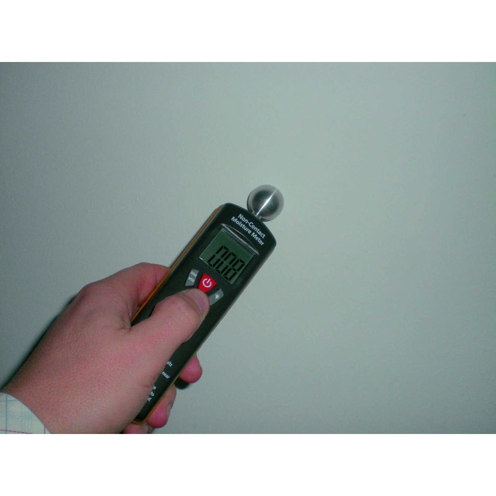 Hand hält ein feuchtigkeitsmessgerät mit digitaler Anzeige vor einer hellen Wand.