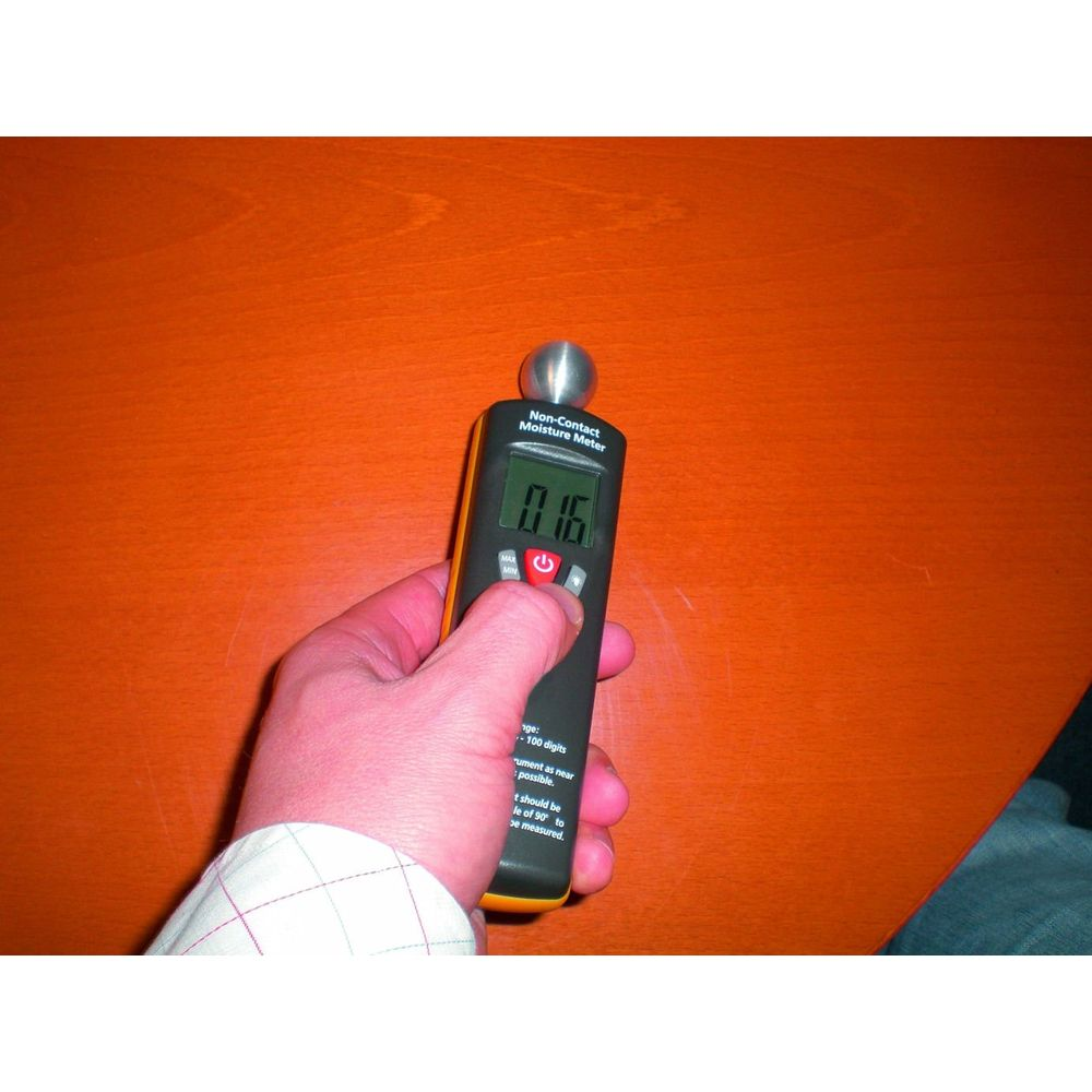Eine Hand hält ein digitales Feuchtigkeitsmessgerät auf einer Holztischoberfläche.