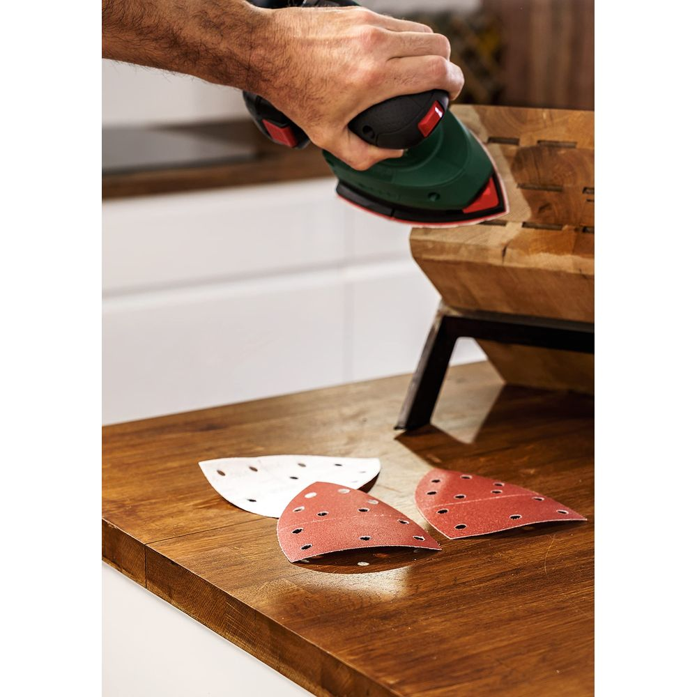 Hand schuurt houten object met groene schuurmachine. Drie schuurpapieren liggen op tafel.
