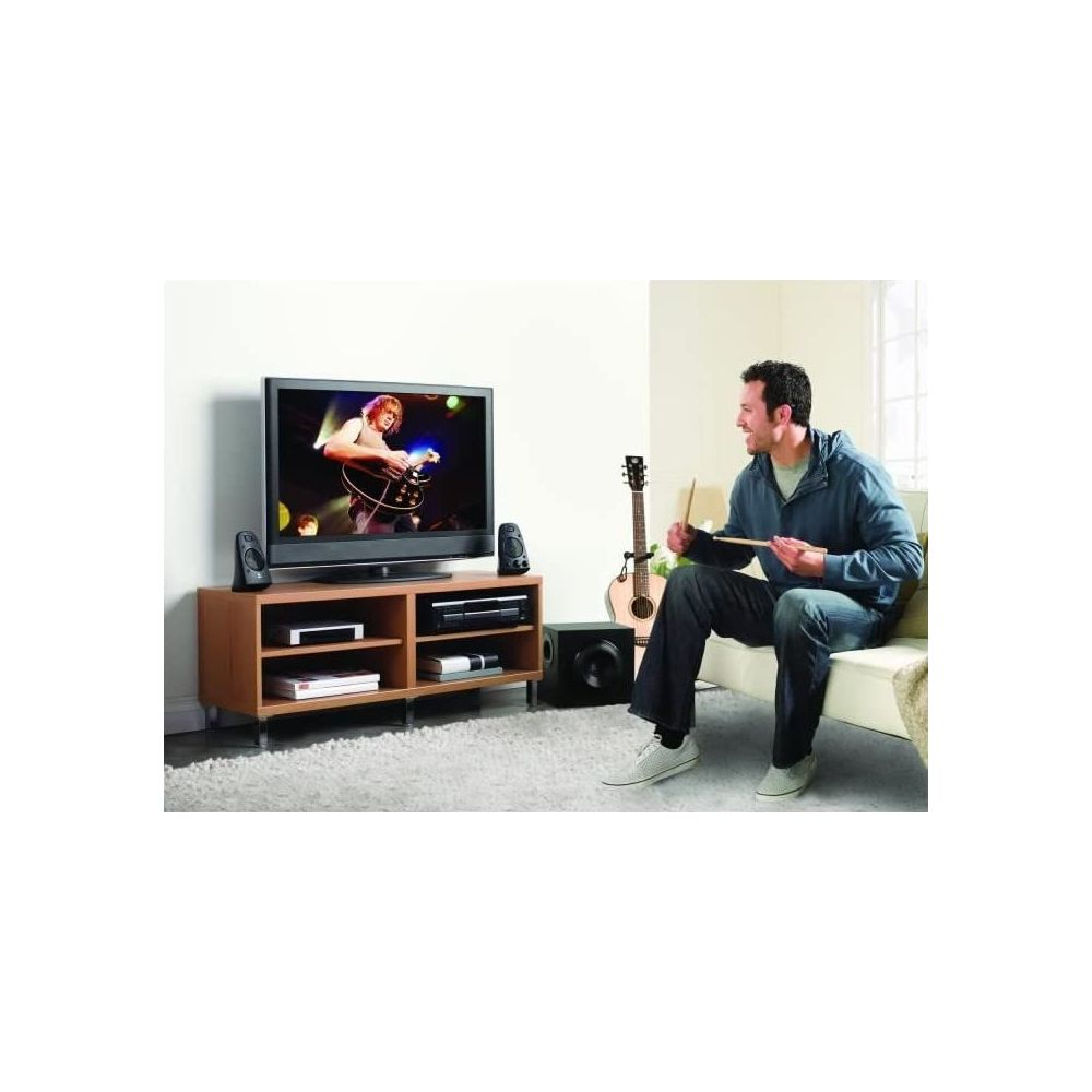 Mężczyzna w niebieskiej bluzie z kapturem i dżinsach siedzi na białej kanapie, trzymając pałeczki, oglądając telewizor z gitarzystą.