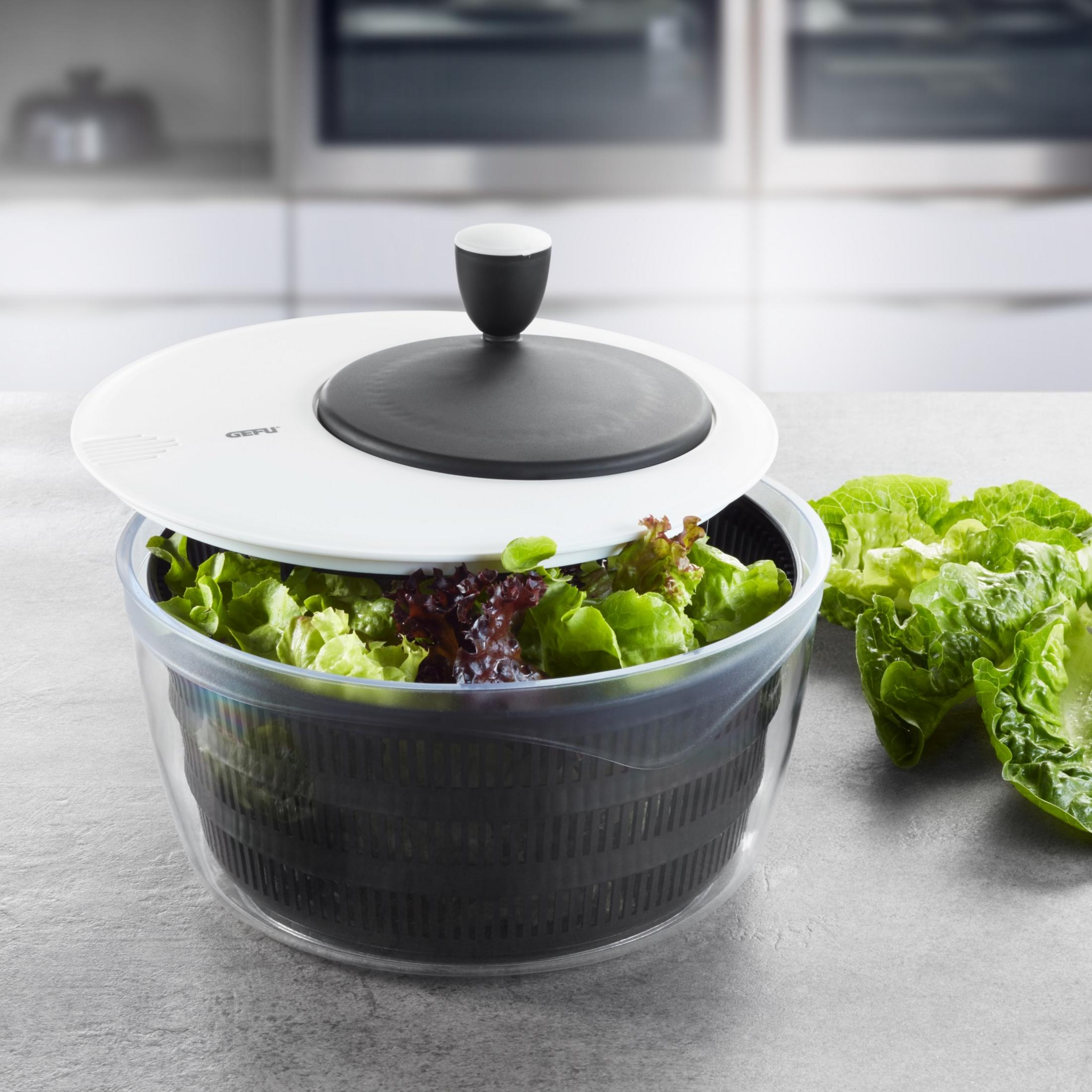 Salatschleuder mit Salat gefüllt, daneben liegen Salatblätter auf grauem Untergrund.