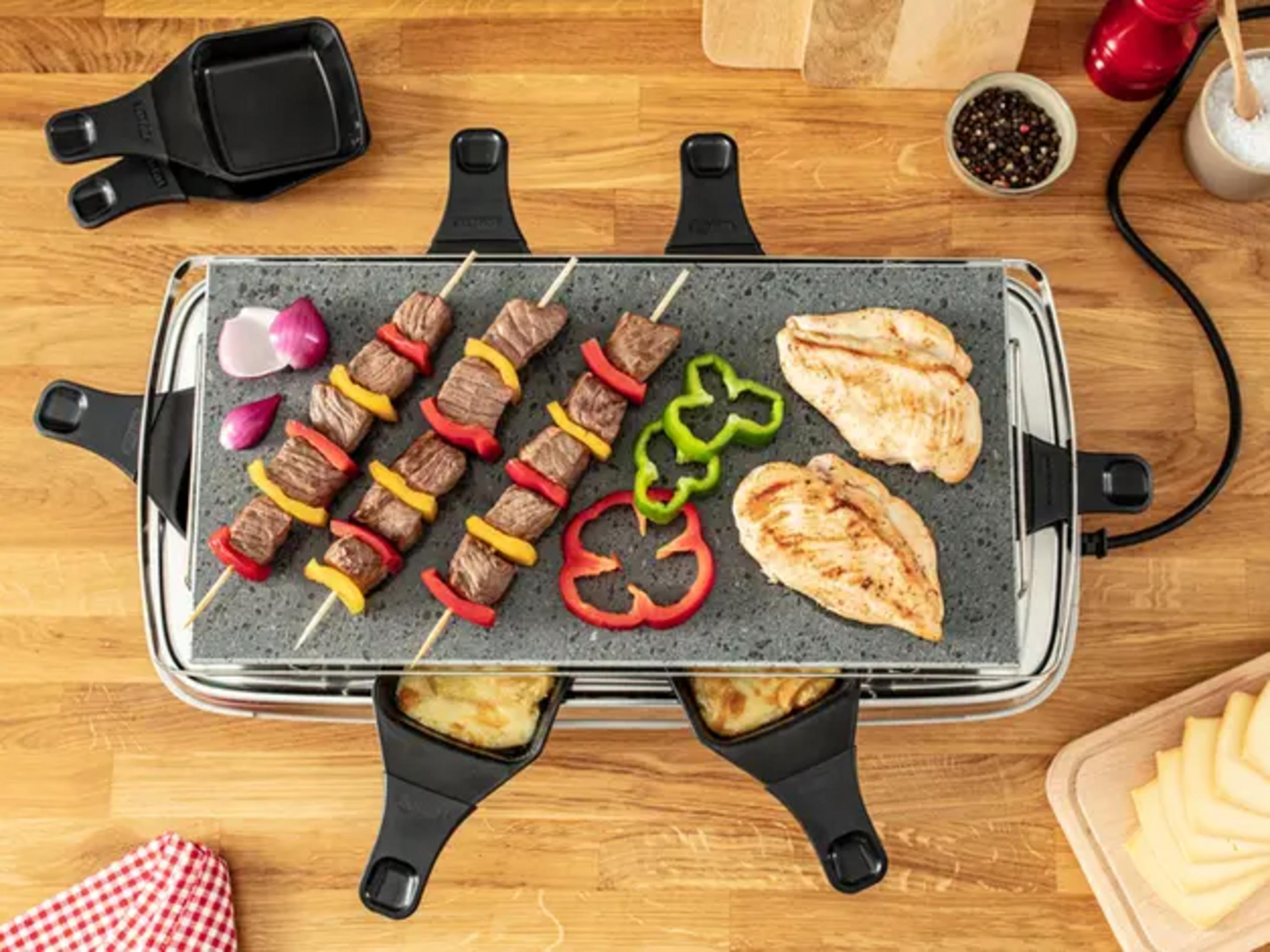 Raclette mit Fleischspießen, Hähnchenbrust und Käse auf einem Holztisch.
