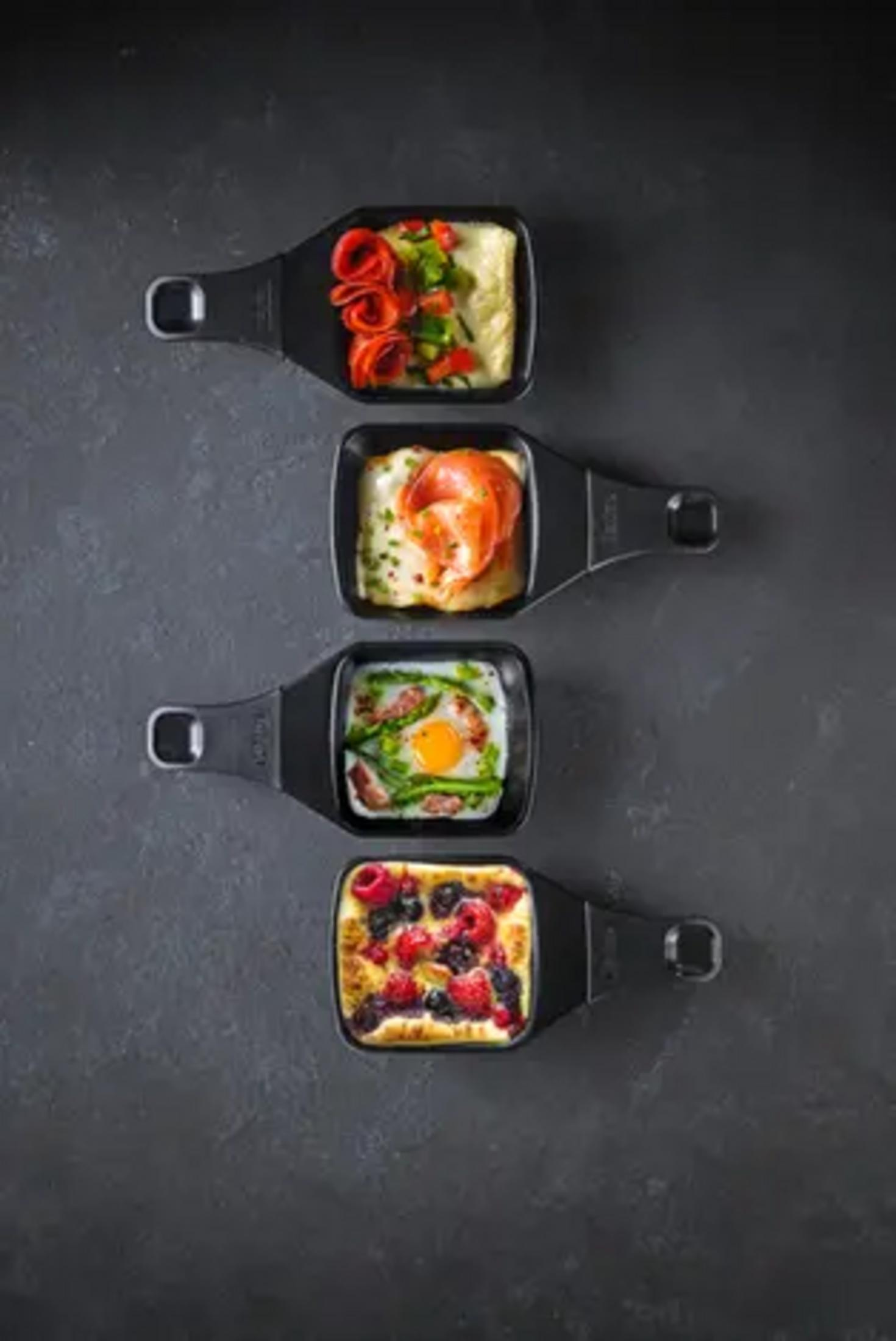 Vier Raclette-Pfännchen mit verschiedenen Speisen: Beeren-Auflauf, Ei mit Spargel, Lachs und Gemüse-Auflauf.