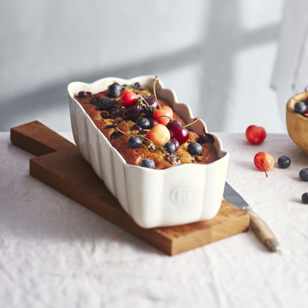 Ein Kuchen mit Beeren und Kirschen in einer weißen Form auf einem Holzbrett.
