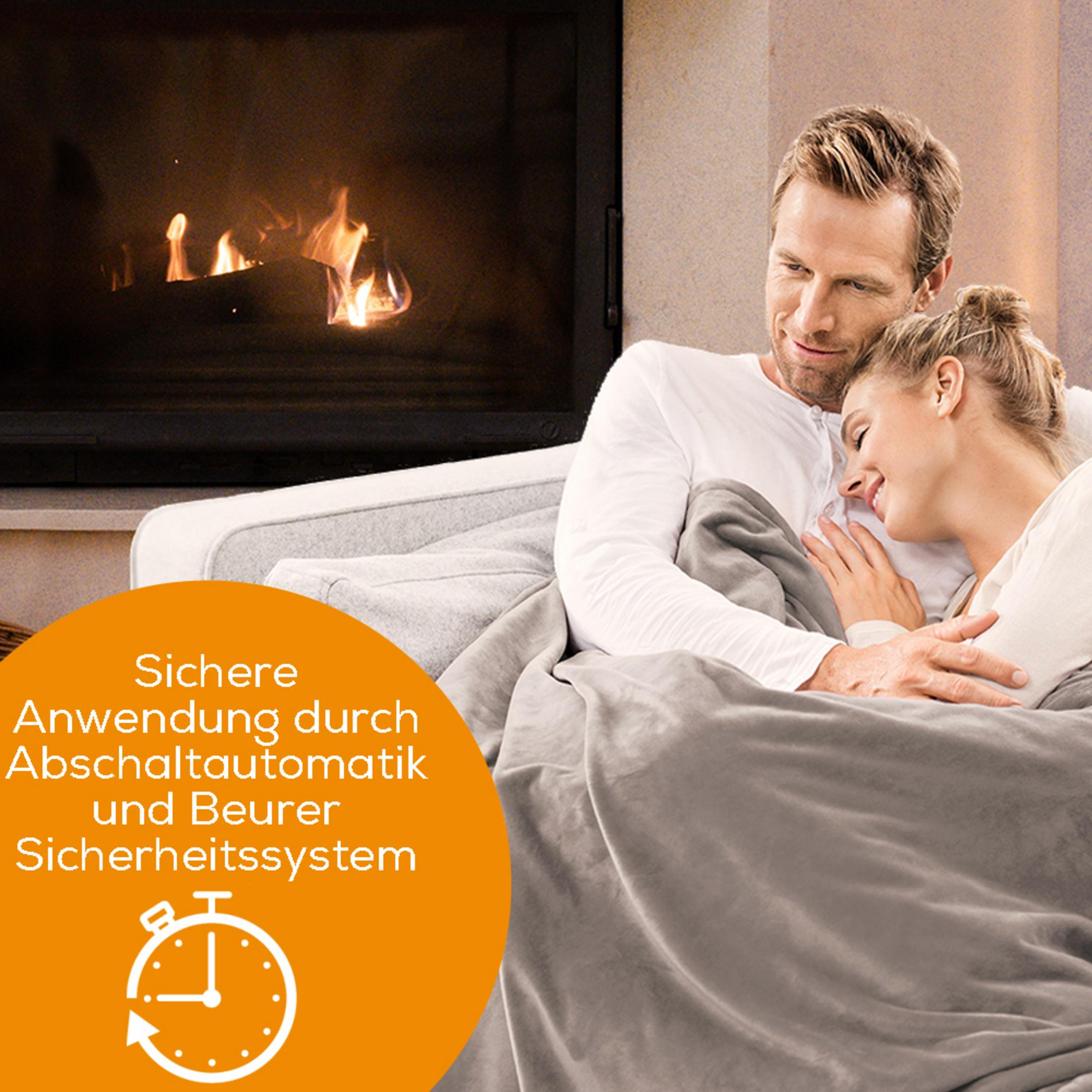 A couple cuddles under a blanket by a fireplace.
