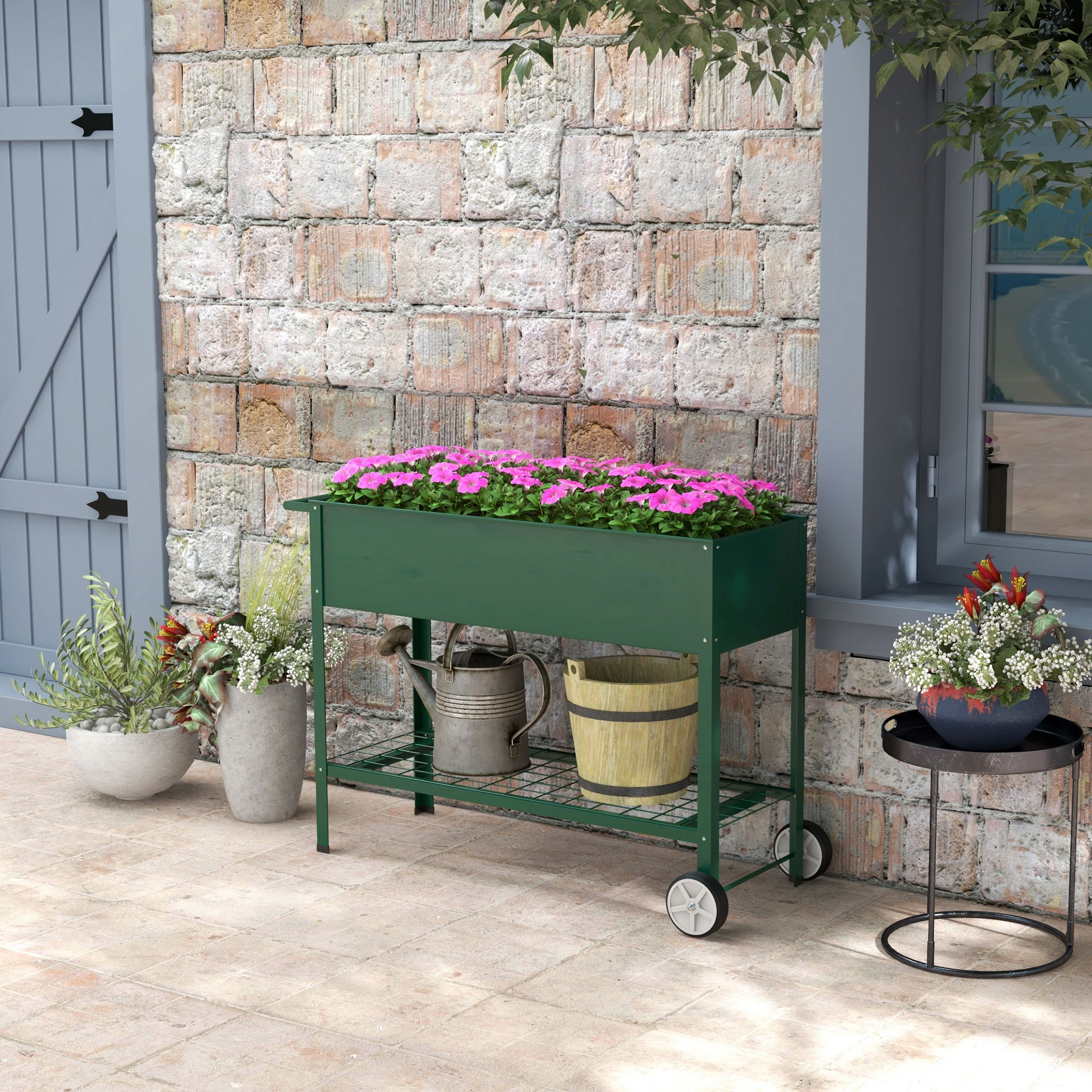 Ein grüner Blumenkasten mit rosa Blumen auf Rädern, daneben eine Gießkanne und ein Holzeimer.