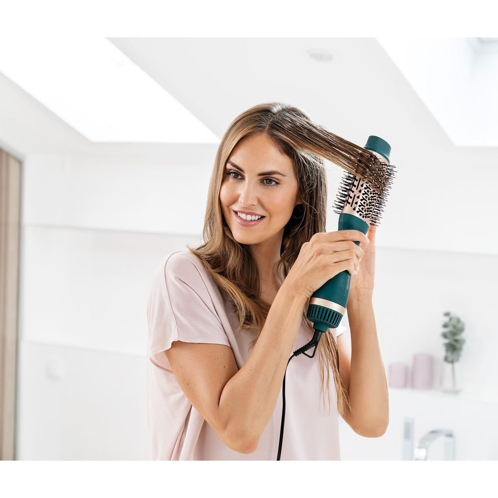 Una mujer se seca el pelo castaño con un cepillo secador giratorio azul verdoso.